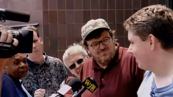 Michael Moore in a cap speaking to reporters holding microphones and cameras