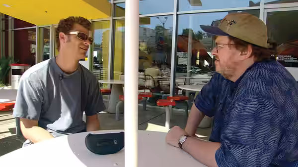 Michael Moore in a cap talking with a younger man at an outdoor table