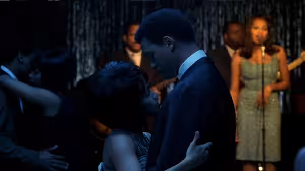 A suited man embraces a woman at a party as a singer performs