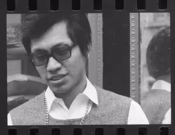 A young man in dark glasses and a sweater vest stands beside a mirror