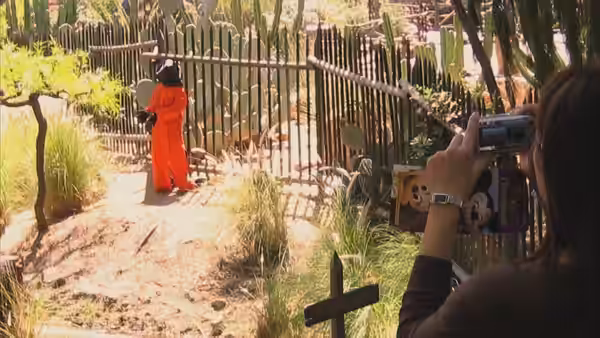 A person in an orange jumpsuit stands by a fence while another films outdoors