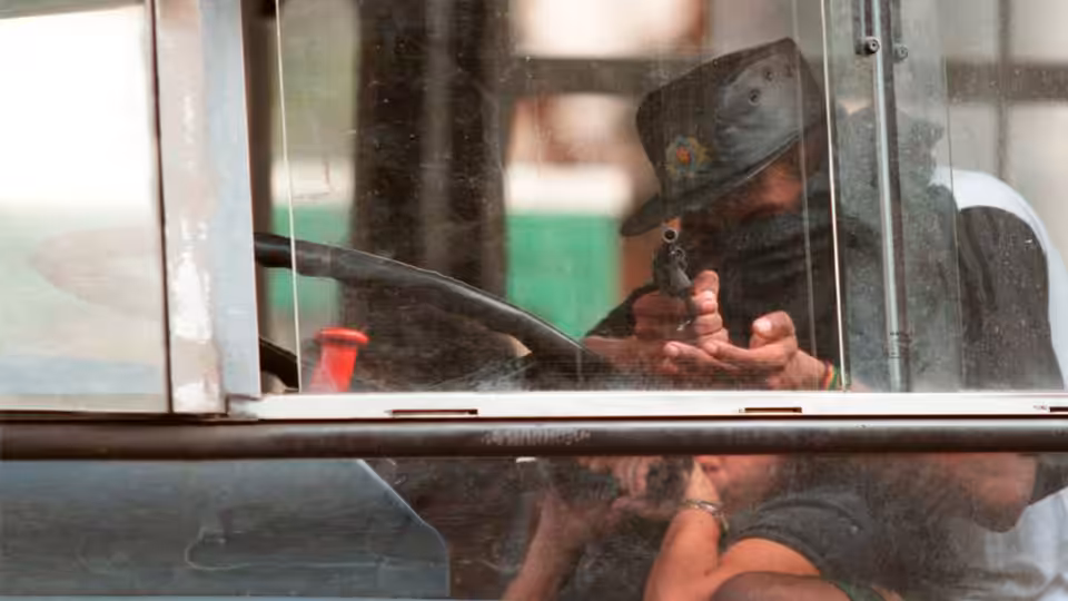 A man aims a handgun through a bus window as passengers crouch below