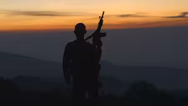 Silhouetted armed figure stands before hills under an orange sunset