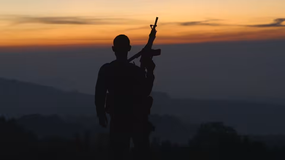Silhouetted armed figure stands before hills under an orange sunset
