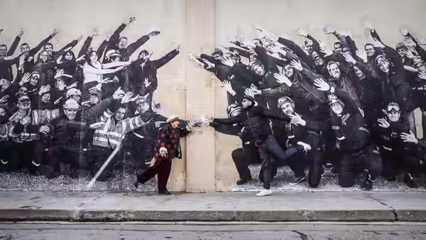Agnès Varda (Agnès Varda) leans against a wall with a reaching crowd mural