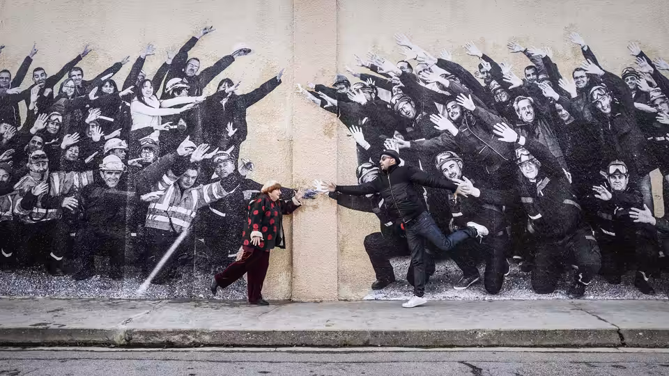 Agnès Varda (Agnès Varda) leans against a wall with a reaching crowd mural