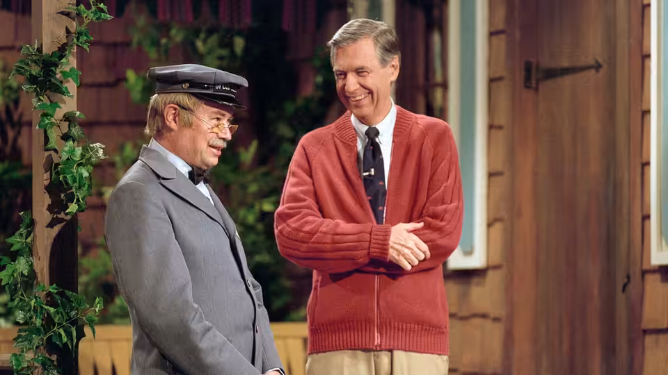 Fred Rogers in red cardigan stands with Mr. McFeely outside a house