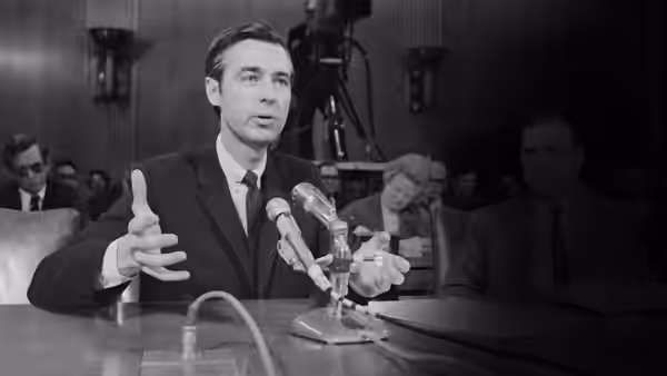 Man in dark suit speaks into microphones at a hearing table