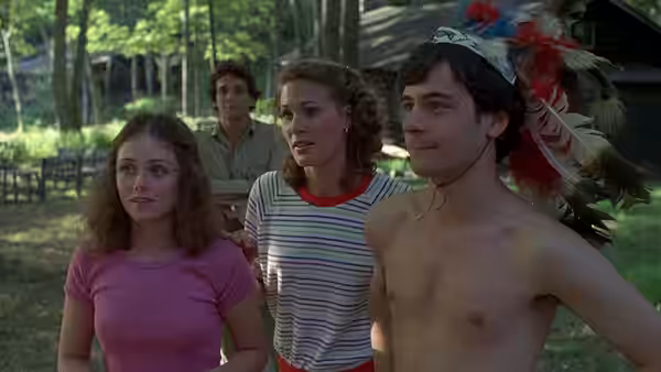 Three camp counselors stand together outside a cabin, one wearing a feathered headdress
