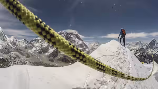 A mountaineer stands on a snowy ridge beside a climbing rope, with peaks behind him