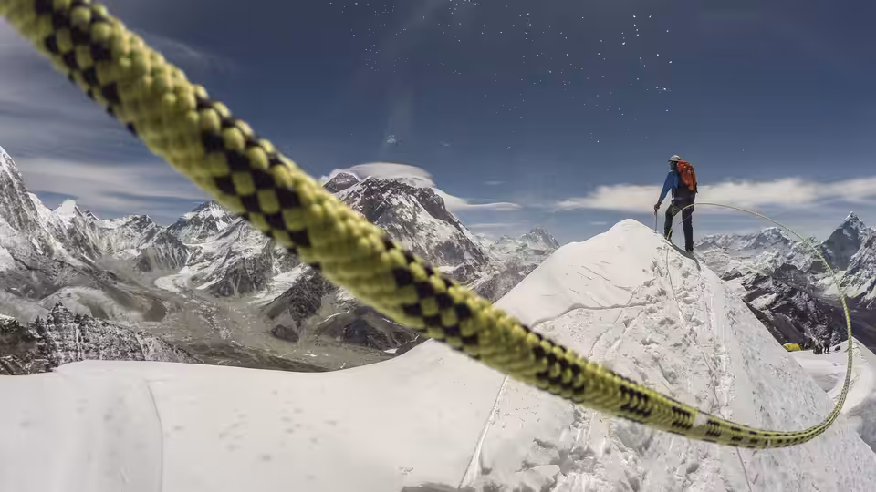 A mountaineer stands on a snowy ridge beside a climbing rope, with peaks behind him
