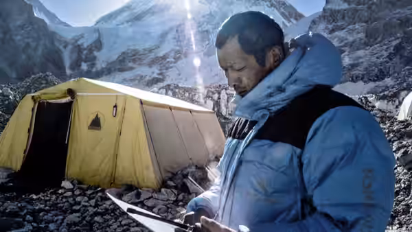 A man in a blue down jacket checks equipment beside a tent in a rocky mountain camp
