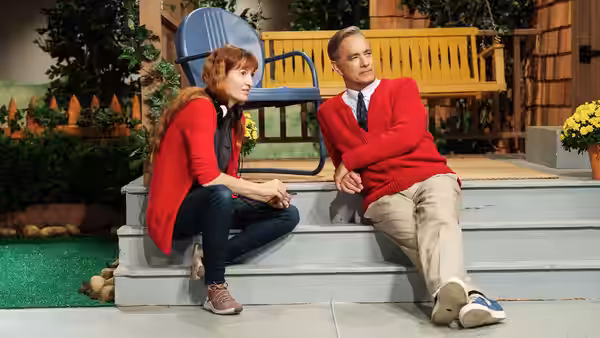 Fred Rogers (Tom Hanks) sits on steps beside a red-haired woman on the set