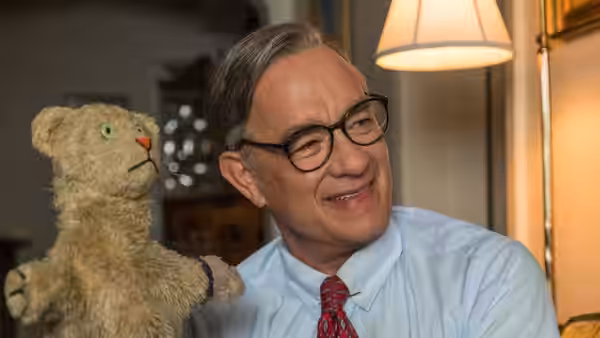 Fred Rogers (Tom Hanks) smiles beside a puppet in a warmly lit room