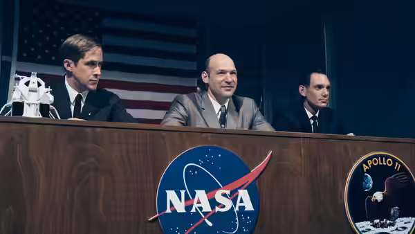 Three men seated at a desk during a NASA press conference with flag behind