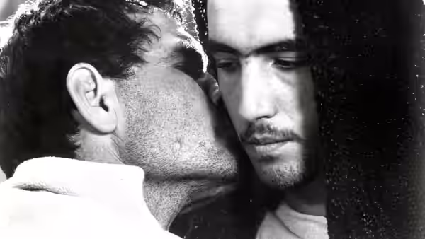 A bearded man whispers into Cristo's ear as Cristo looks down