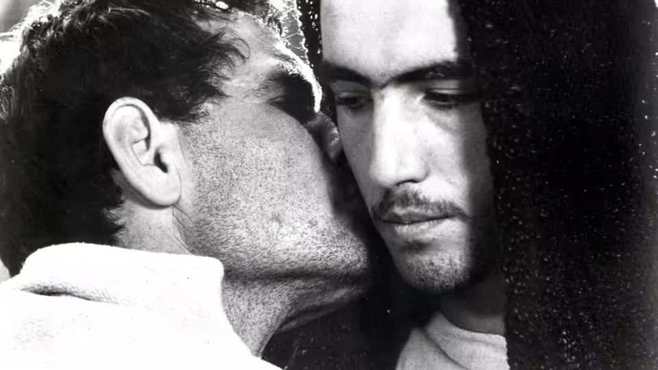 A bearded man whispers into Cristo's ear as Cristo looks down