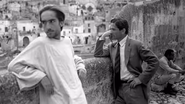Cristo stands beside a suited man on a terrace overlooking a town