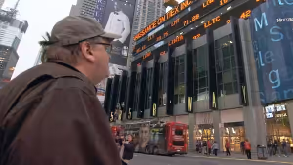 Michael Moore looks up at a building covered in stock market tickers