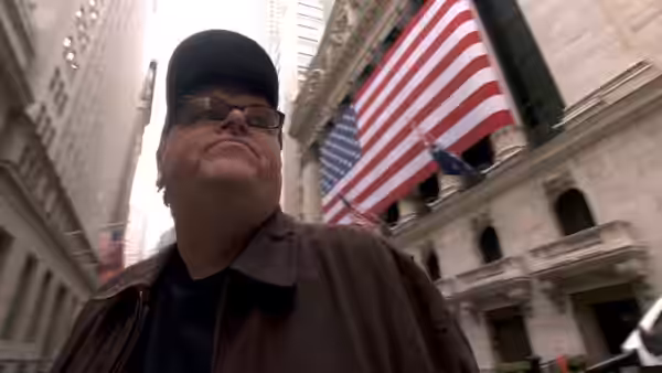 Michael Moore in a cap and glasses stands beneath a large American flag