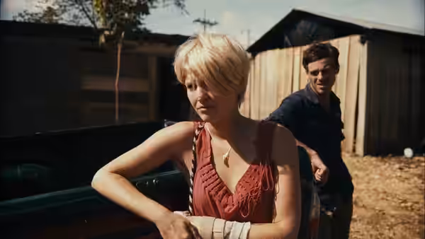 Samantha (Whitney Able) leaning on a car with Andrew (Scoot McNairy) behind her