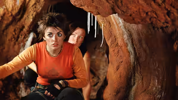 Woman in orange shirt crouches alert in a cave opening