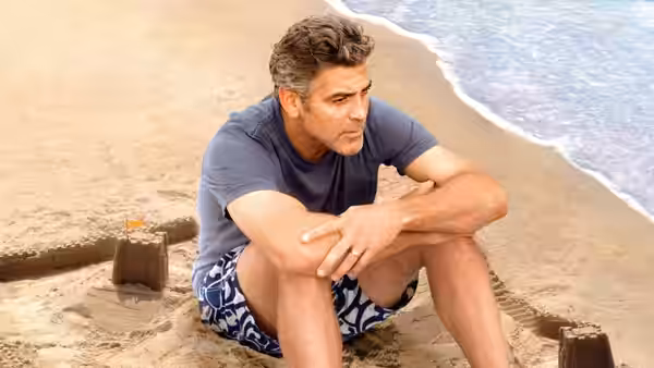 Matt King (George Clooney) sitting on the sand with arms folded near the shoreline