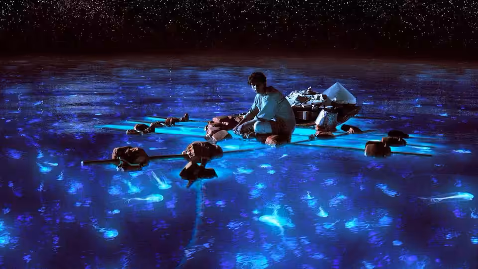 Young man sits cross-legged on raft amid glowing blue sea creatures at night
