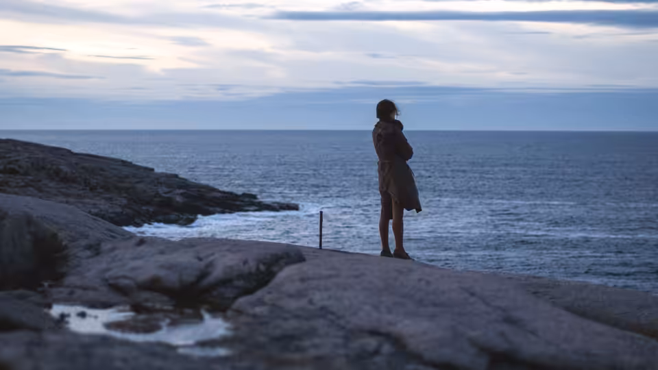 a woman in a brown coat standing on rocks by the sea