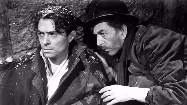 An injured man crouches beside a companion in a wide-brimmed hat