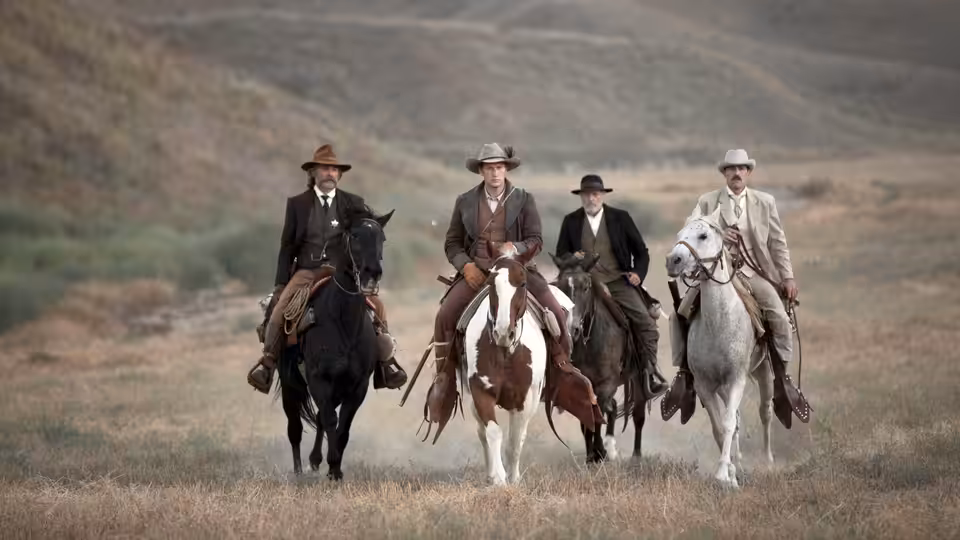 Four men on horseback riding across open grassland in a line