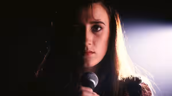 Woman with dark hair sings into a microphone under a spotlight