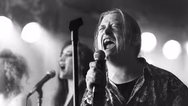 Singer with long hair shouts into a microphone on a brightly lit stage