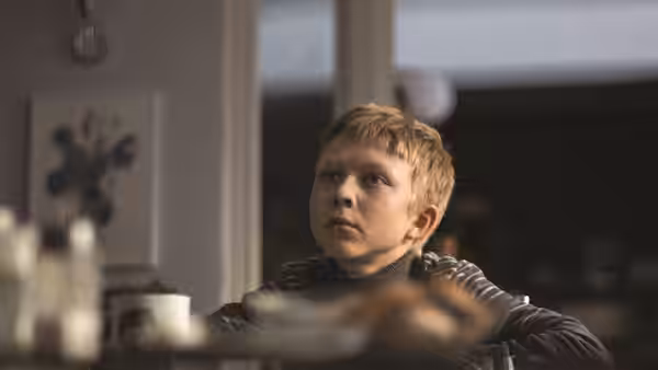 Alyosha (Matvey Novikov) sits at table looking up with neutral expression