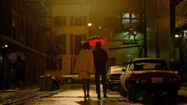 A couple walks arm in arm under a red umbrella on a wet city street at night
