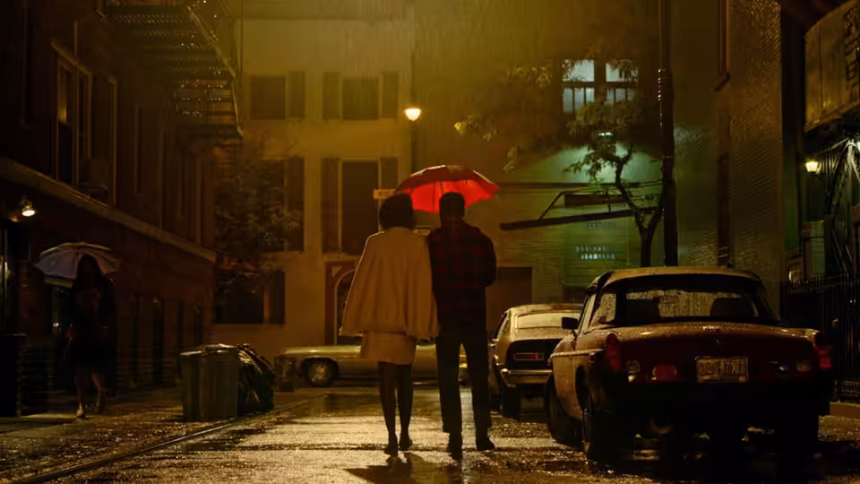 A couple walks arm in arm under a red umbrella on a wet city street at night