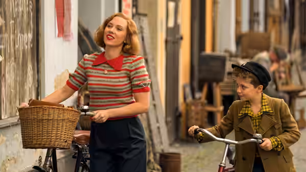 Rosie (Scarlett Johansson) walks beside Jojo (Roman Griffin Davis), who rides a bicycle down a street