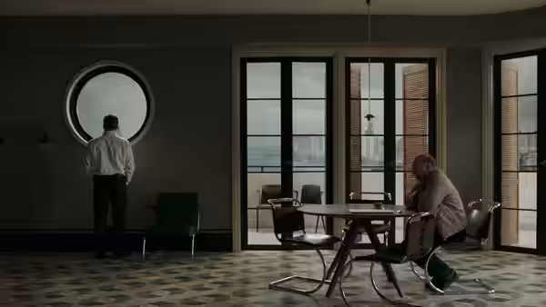 one man stands at a round window while another sits at a table