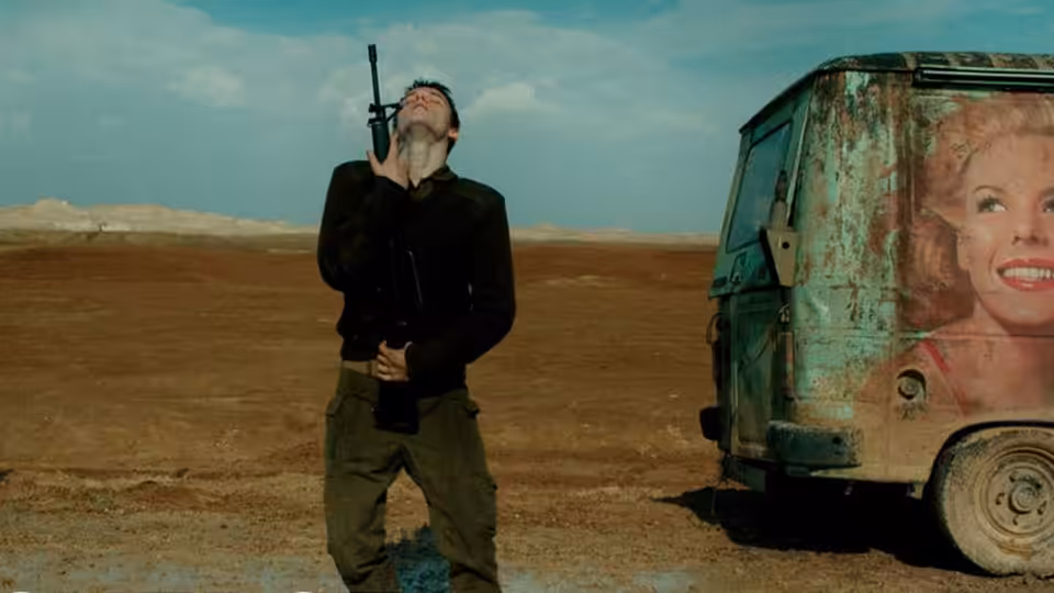 a bearded man holds a rifle beside a rusted van in the desert