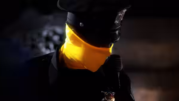 Police officer wearing black uniform and yellow neck gaiter holding a radio at night