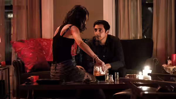 Nasir Khan (Riz Ahmed) seated on a couch while a woman leans over the table toward him