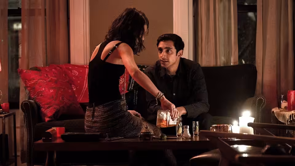 Nasir Khan (Riz Ahmed) seated on a couch while a woman leans over the table toward him