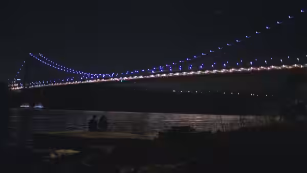 Two figures sit on a pier beneath a bridge illuminated with colorful lights at night