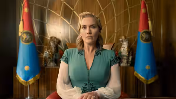 Chancellor Elena Vernham (Kate Winslet) seated in a red chair between two flags