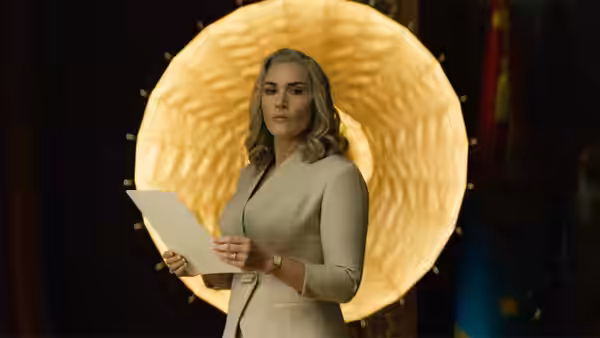 Chancellor Elena Vernham (Kate Winslet) holding papers before a large golden light