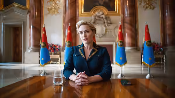 Chancellor Elena Vernham (Kate Winslet) seated at a polished table with hands clasped