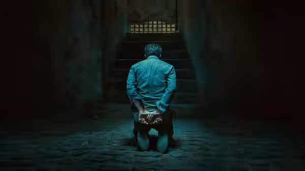 A man in a blue shirt kneels with hands cuffed on a stone floor