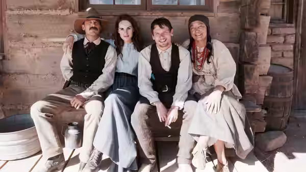 four people sit together on the steps outside a wooden frontier building