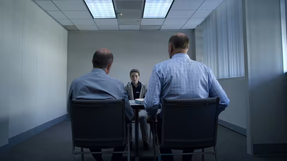 Marie Adler (Kaitlyn Dever) sits facing two men at a table in a small room