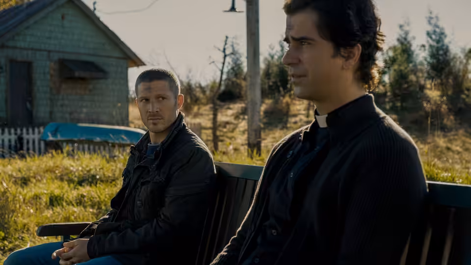 Riley Flynn (Zach Gilford) and Father Paul Hill (Hamish Linklater) seated on a bench outside